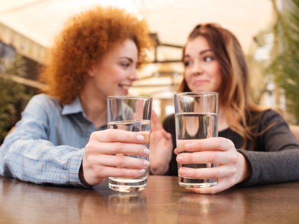 Two smiling charming young women drinking water at the table in cafe