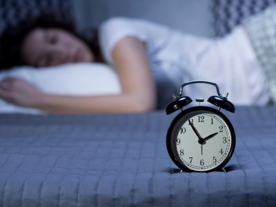Close-up of a black, vintage alarm clock on a bed where a young woman is lying in the blurry background