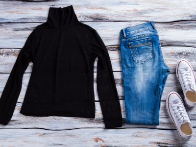 Black top with blue jeans. White canvas shoes and pants. Outfit with high collar top. Female clothing on wooden table.