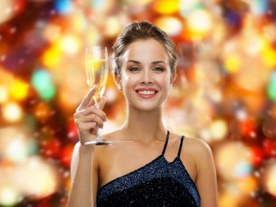 drinks, holidays, christmas, people and celebration concept - smiling woman in evening dress with glass of sparkling wine over red christmas lights background